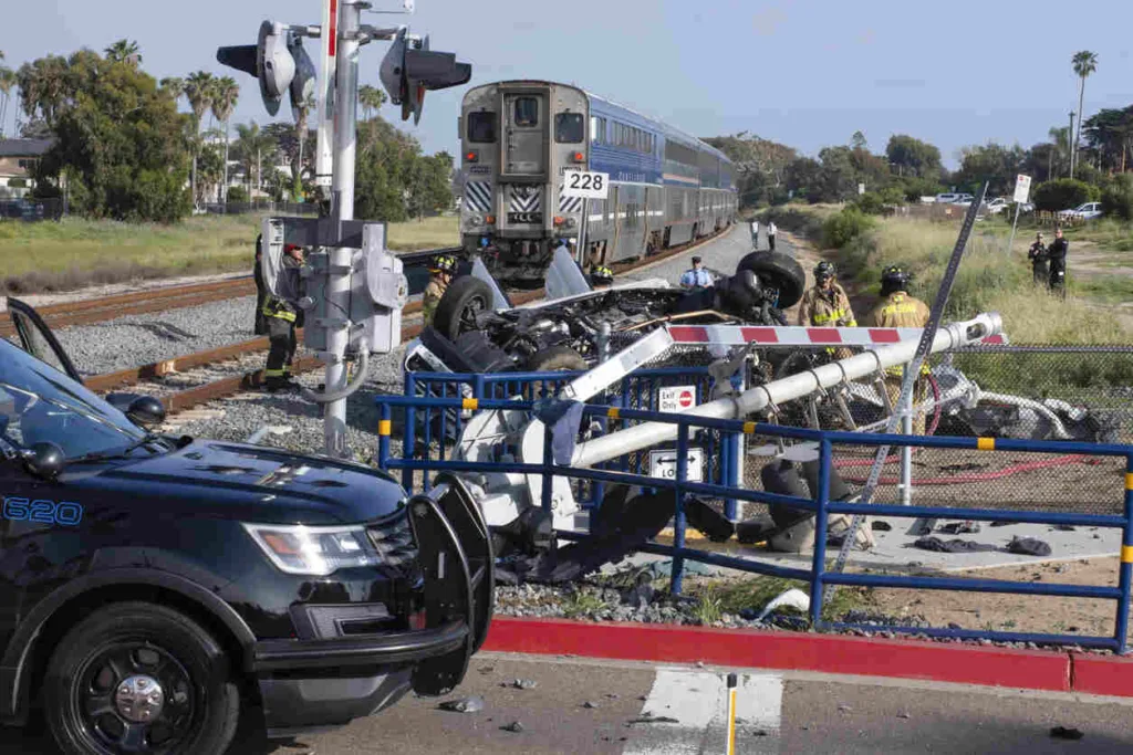 Overturned vehicle at railroad crossing after collision with train, emergency responders and police on scene, train stopped on tracks, clear daylight