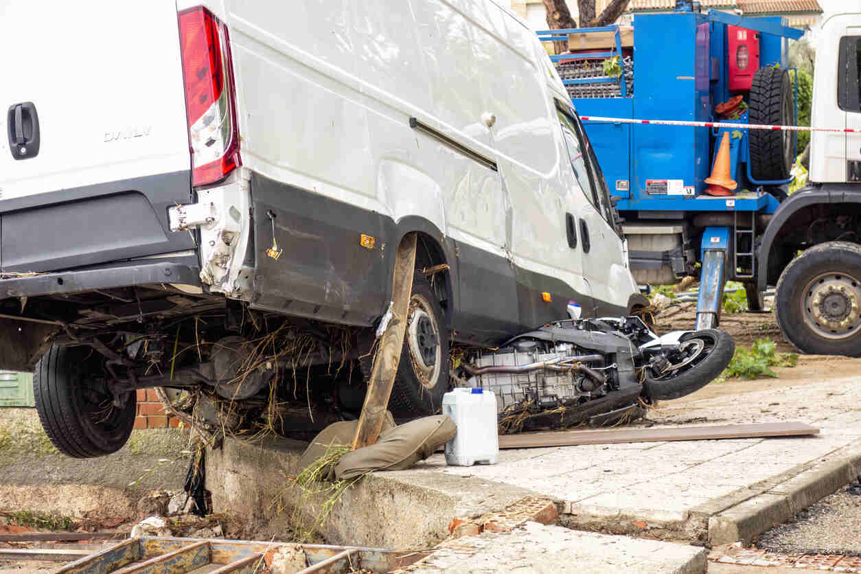 A damaged delivery van rests precariously on a roadside barrier after a collision with a motorcycle, suggesting a potential lost load incident.