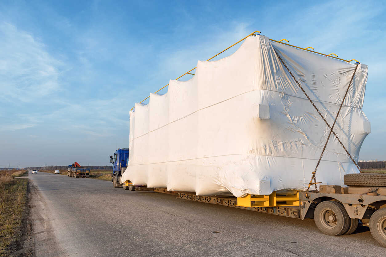An oversized load, wrapped in a white tarp and secured with chains, is being hauled on a flatbed semi-truck on a highway, illustrating the potential for oversized load accidents.