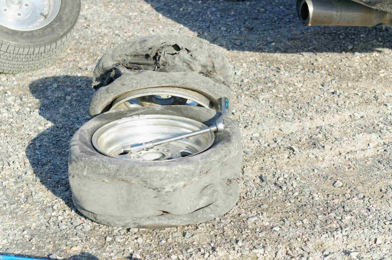 A severely damaged, blown-out tire lies on a gravel surface next to a vehicle, illustrating the aftermath of a tire blowout accident.